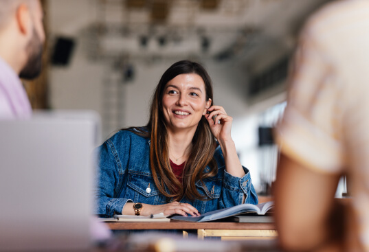 A woman sitting with a book open in front of her. She is sitting across from two other people smiling.